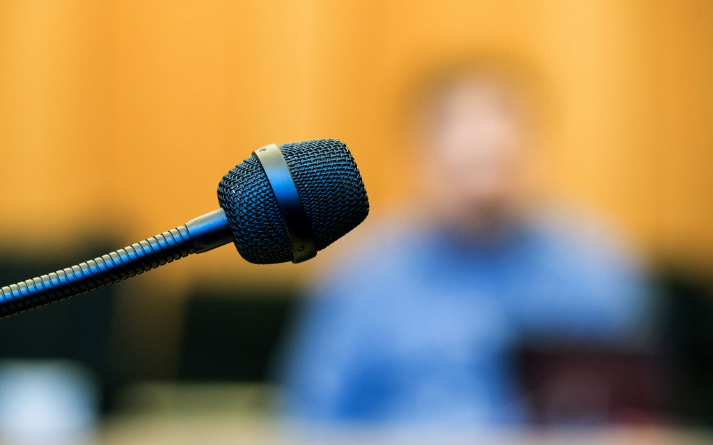 A blurred image of a witness behind a microphone in a courtroom